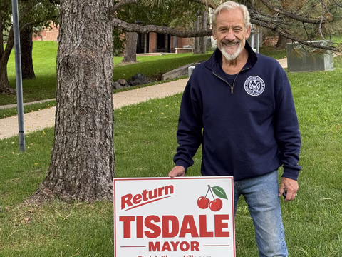 Doug Tisdale standing beside a campaign yard sign beneath mature trees.